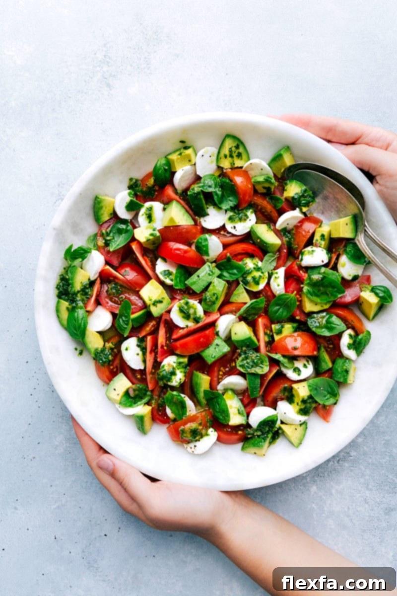 A vibrant Caprese Salad in a bowl, featuring juicy red tomatoes, creamy white mozzarella pearls, lush green basil leaves, and sliced avocado, all glistening with a homemade basil vinaigrette. Fresh torn basil leaves are scattered on top as a garnish.