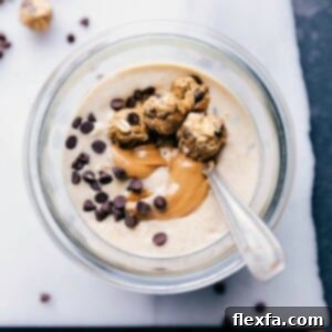 Close-up of a single serving of Peanut Butter Cookie Overnight Oats in a clear jar, garnished with a swirl of peanut butter and topped with mini no-bake cookies and chocolate chips.