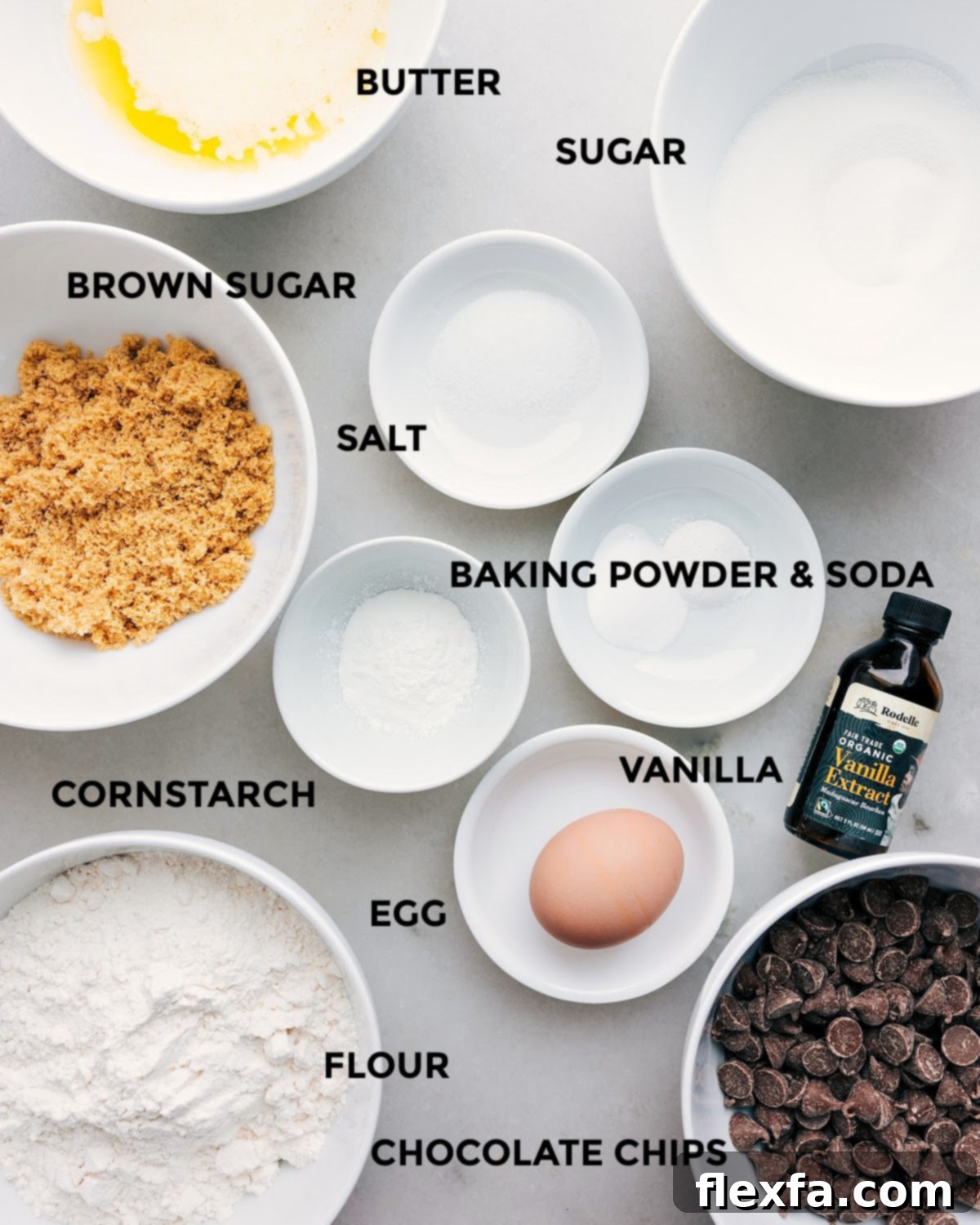 All the essential ingredients for bakery-style chocolate chip cookies laid out on a clean surface, including butter, various sugars, egg, vanilla, cornstarch, leavening agents, salt, flour, and a generous bowl of chocolate chips.