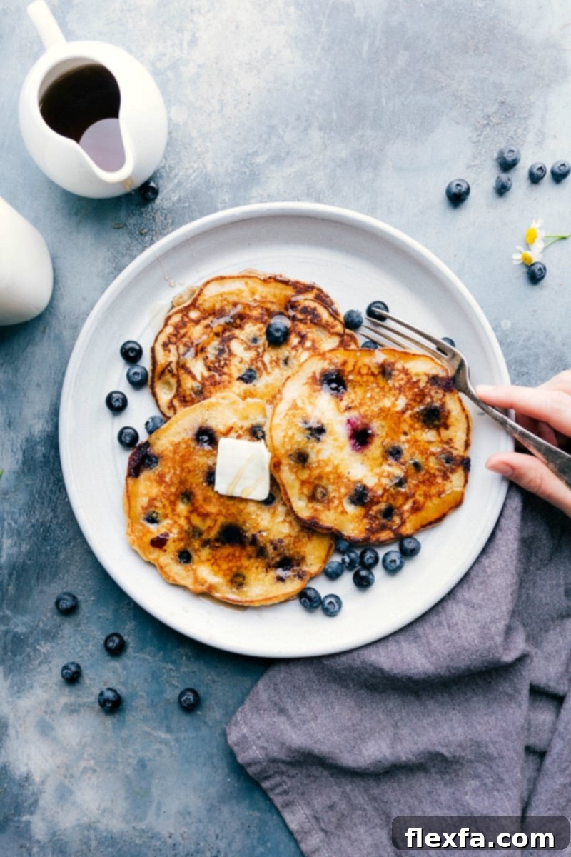 A stack of warm blueberry pancakes on a plate, topped with melting butter and drizzled with syrup, with a fork on the side ready for enjoying.
