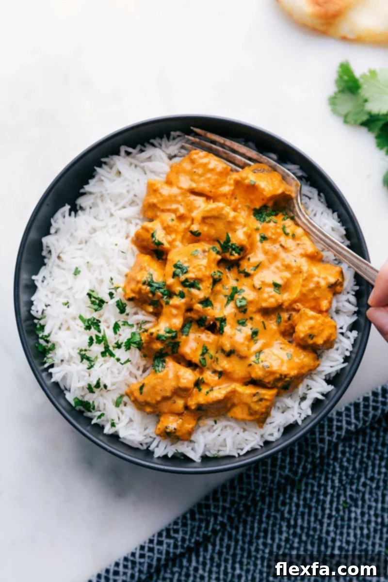 A steaming bowl of homemade Chicken Tikka Masala served with fluffy Basmati rice, garnished with fresh cilantro. A fork gently scoops up a piece of succulent chicken from the creamy, orange-hued sauce, highlighting its tender texture and the rich consistency of the curry.