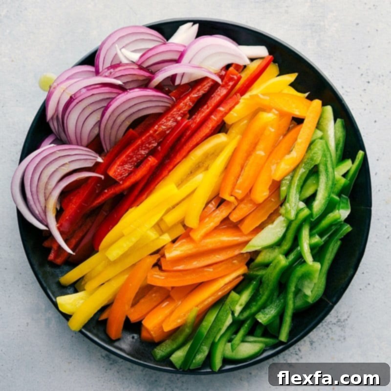 A colorful array of sliced bell peppers in red, orange, yellow, and green, alongside thinly sliced red onion, meticulously prepared for a sheet pan fajita recipe.