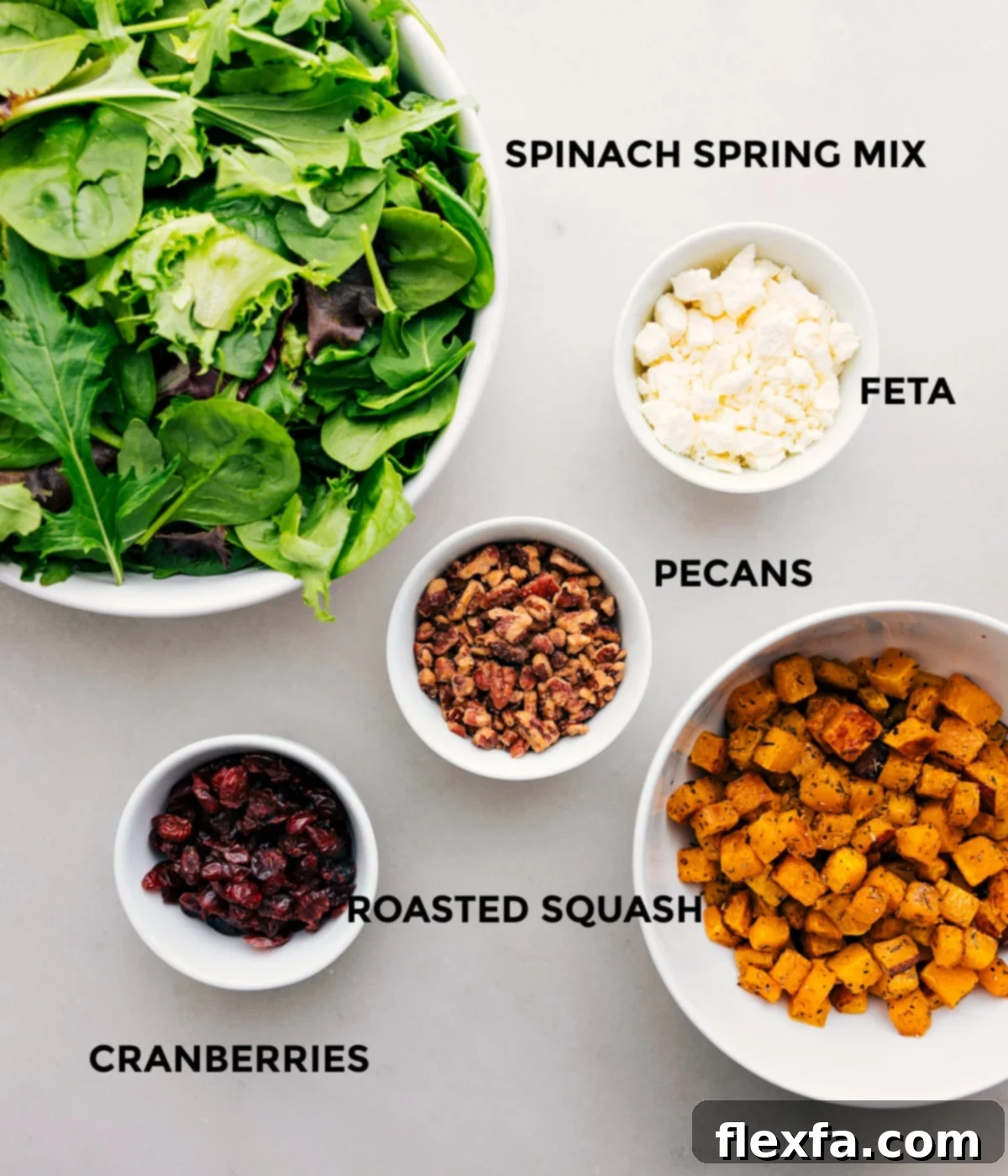 Essential Ingredients For A Delicious Thanksgiving Salad A collection of fresh, vibrant ingredients for Thanksgiving Salad, including leafy greens, crumbled feta cheese, shiny candied pecans, roasted butternut squash cubes, and dried cranberries, neatly arranged and ready for mixing.