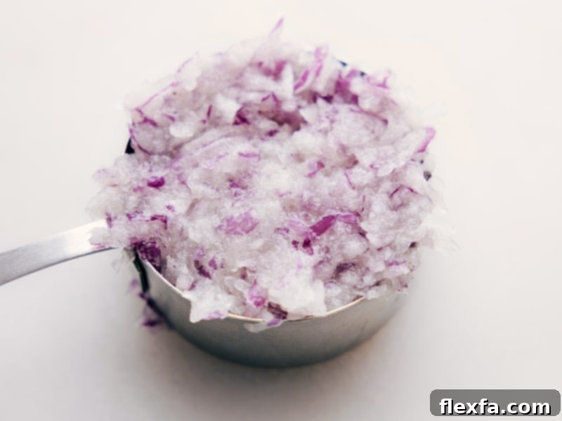 A measuring cup filled with finely grated red onion, showcasing the precise preparation needed for optimal flavor and texture in Greek Meatballs.