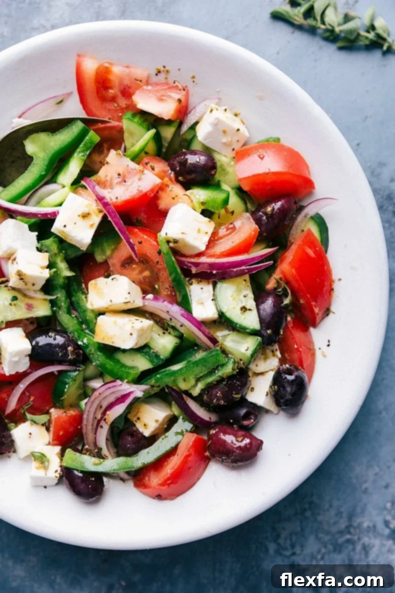 Fresh greek salad in a bowl with tomatoes, olives, feta cheese, and cucumbers. A bottle of olive oil and lemons are also visible on a white tabletop, suggesting healthy ingredients.