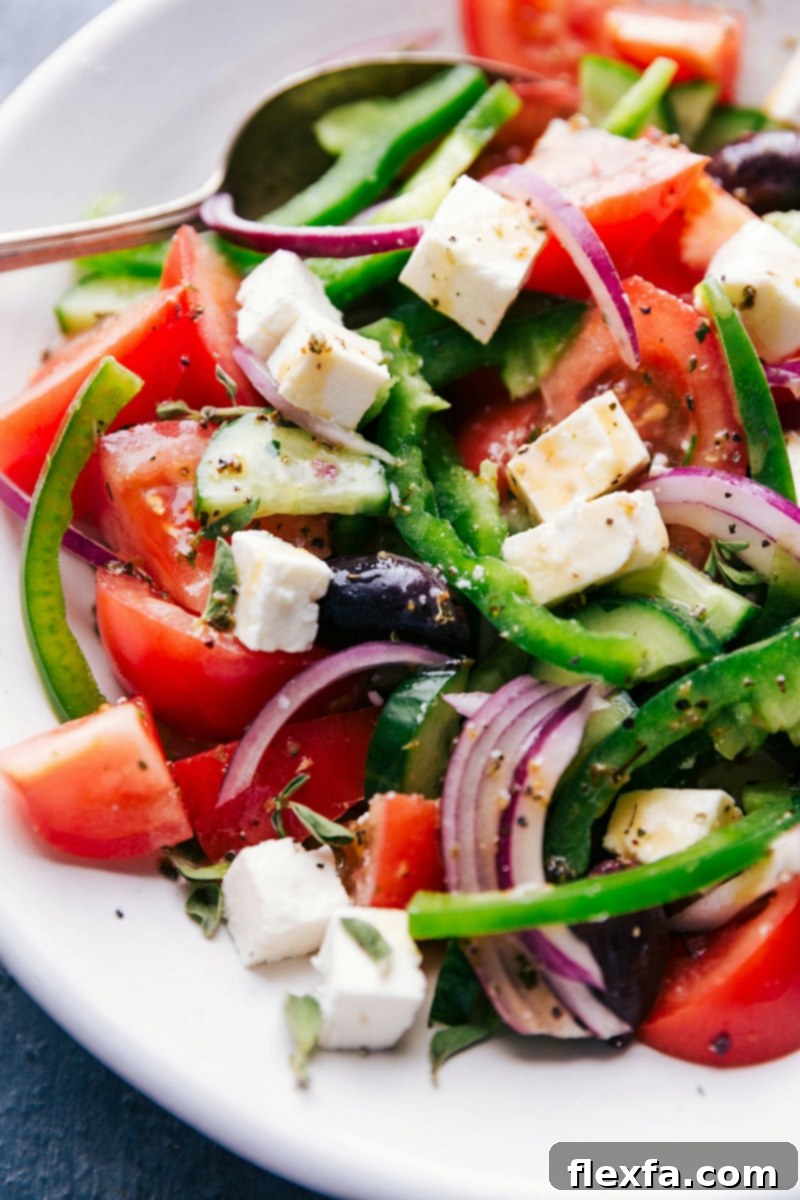 Completed greek salad recipe drizzled in dressing, ready to serve in a large bowl. Freshly chopped tomatoes, cucumbers, feta, and olives are visible.