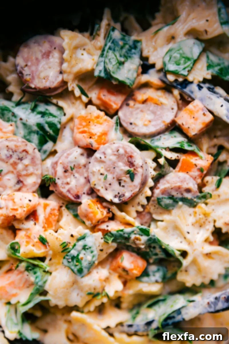 An indulgent Sweet Potato Pasta with roasted and seasoned sweet potatoes, onions, and chicken sausage. The pasta and veggies are tossed in an unforgettable creamy goat cheese sauce and topped with Parmesan. A quick, easy, and simple dinner recipe featuring bowtie pasta, sausage, and sweet potato. Overhead image of Sweet Potato Pasta in a bowl, garnished with fresh basil