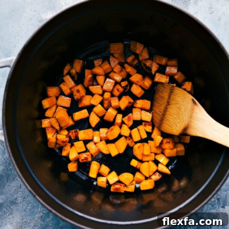 Small, uniformly diced sweet potatoes are sautéing in a large pot with olive oil, softening and beginning to caramelize, preparing for the chili base.