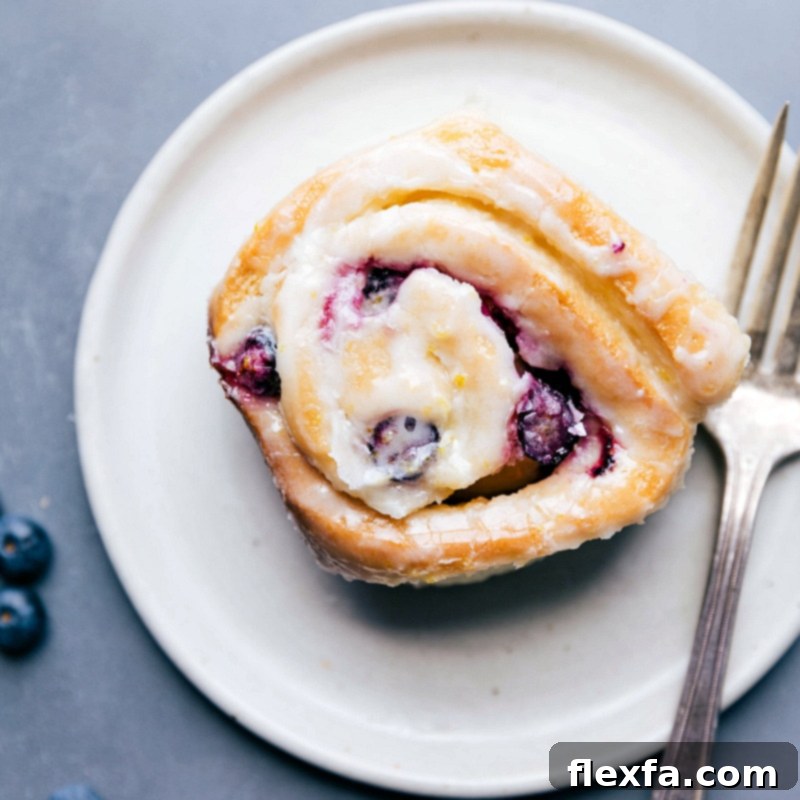 A single, perfectly glazed lemon blueberry sweet roll resting on a white plate, garnished with fresh blueberries and lemon zest, ready to be enjoyed.