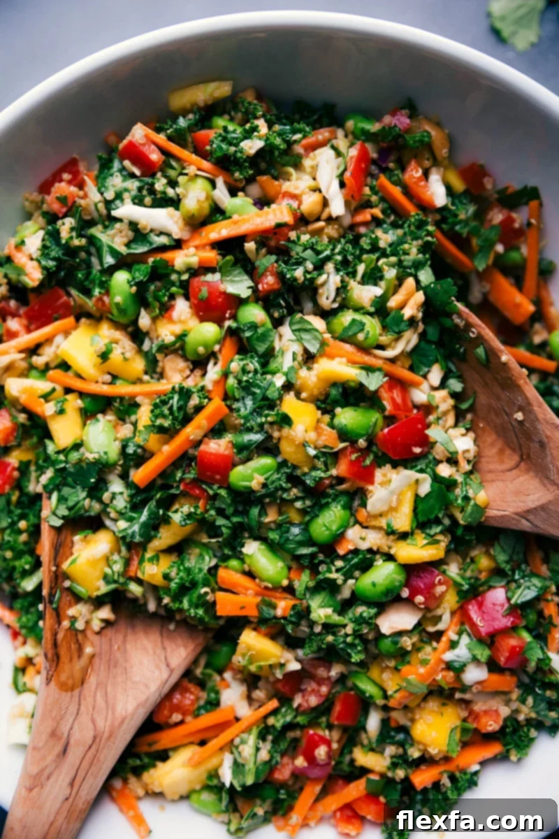 A large, colorful bowl of Thai quinoa salad, featuring fluffy quinoa, crisp red peppers, julienned carrots, bright green edamame, shredded kale, fresh mango chunks, cilantro, and sliced green onions, all generously coated in a rich peanut dressing. It's a healthy, flavorful, and visually appealing dish.