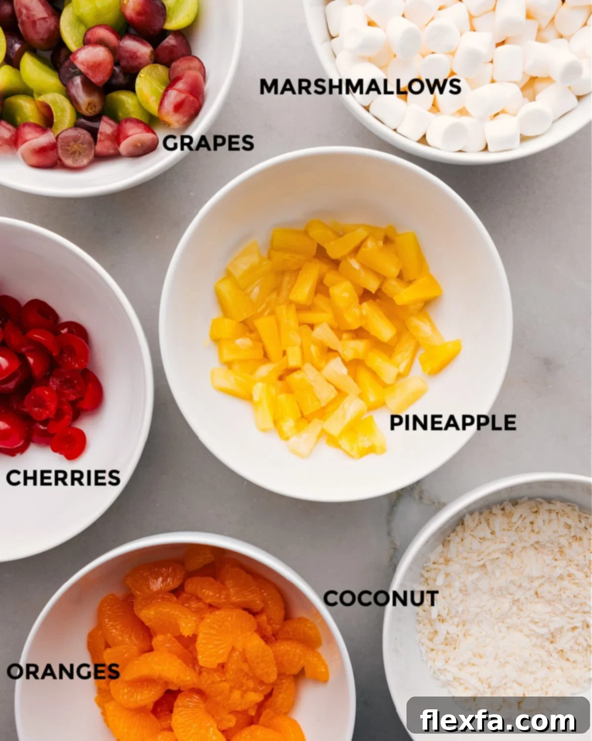 All the fresh and canned ingredients meticulously prepped and laid out for making Ambrosia Salad, including pineapple tidbits, mini marshmallows, green grapes, maraschino cherries, shredded coconut, and mandarin oranges.
