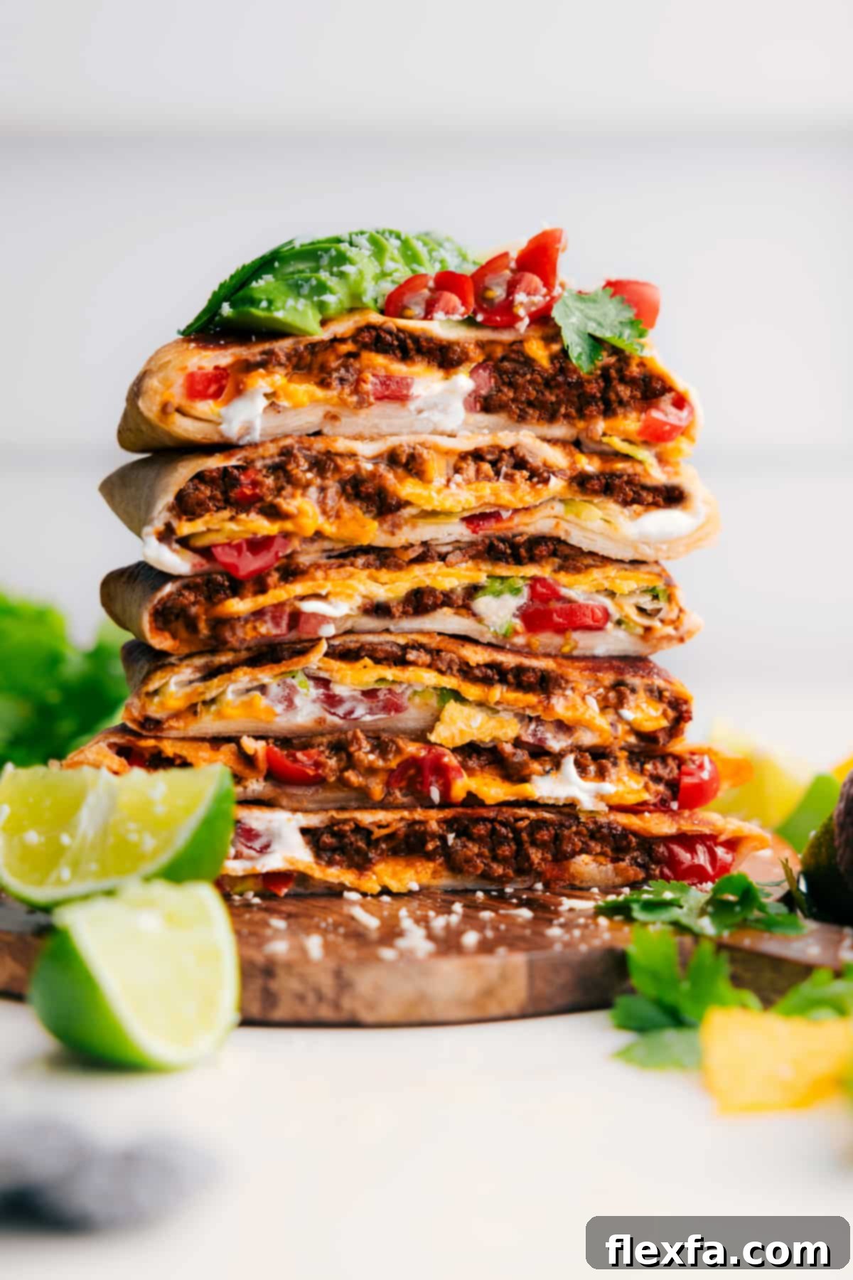 Two halves of a homemade Crunchwrap Supreme, perfectly cut and stacked, revealing the layers of beef, cheese, tostada, and fresh toppings. Garnished with tomatoes and avocado.