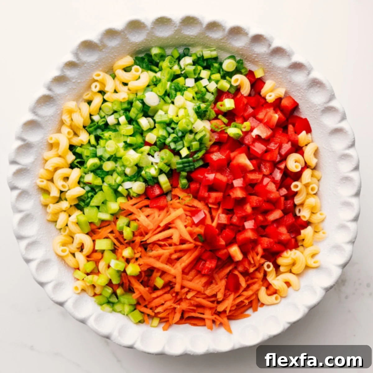 A large bowl containing cooked macaroni, diced celery, red bell pepper, shredded carrots, and sliced green onions, all waiting to be combined with the creamy dressing for macaroni salad.