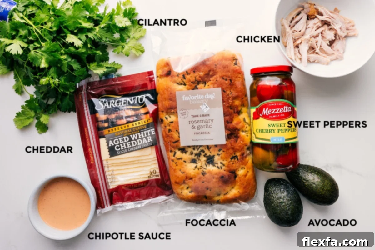 Ingredients for the Panera chipotle chicken melt laid out on a surface, including focaccia bread, shredded chicken, avocado, cilantro, peppers, and chipotle mayo.