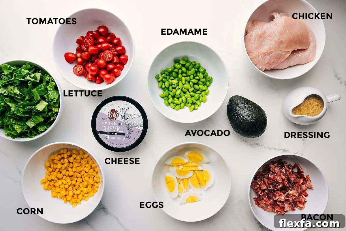 All the ingredients in this recipe prepped out for easy assembly including the lettuce, tomatoes, corn, cheese, edamame, eggs, avocado, dressing, bacon, and chicken.