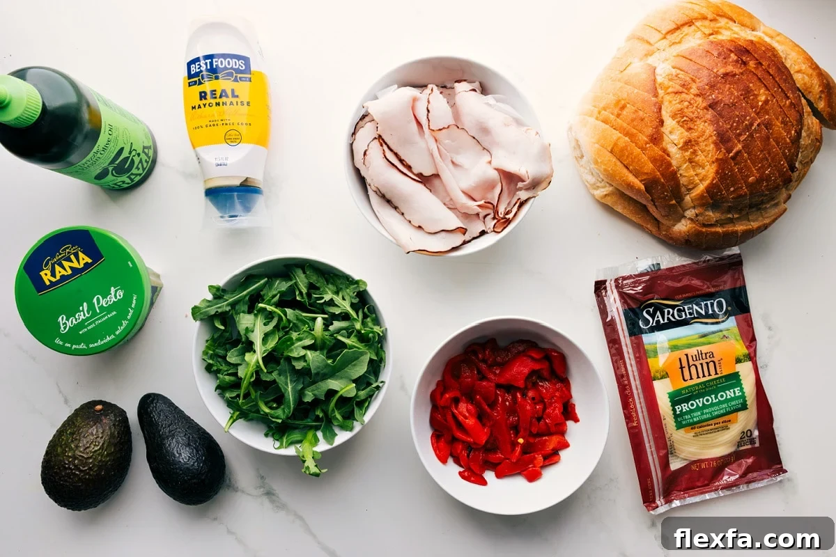 All the ingredients in this recipe prepped out for easy assembly including the oil, pesto, avocado, arugula, turkey, roasted red peppers, provolone, and sourdough.