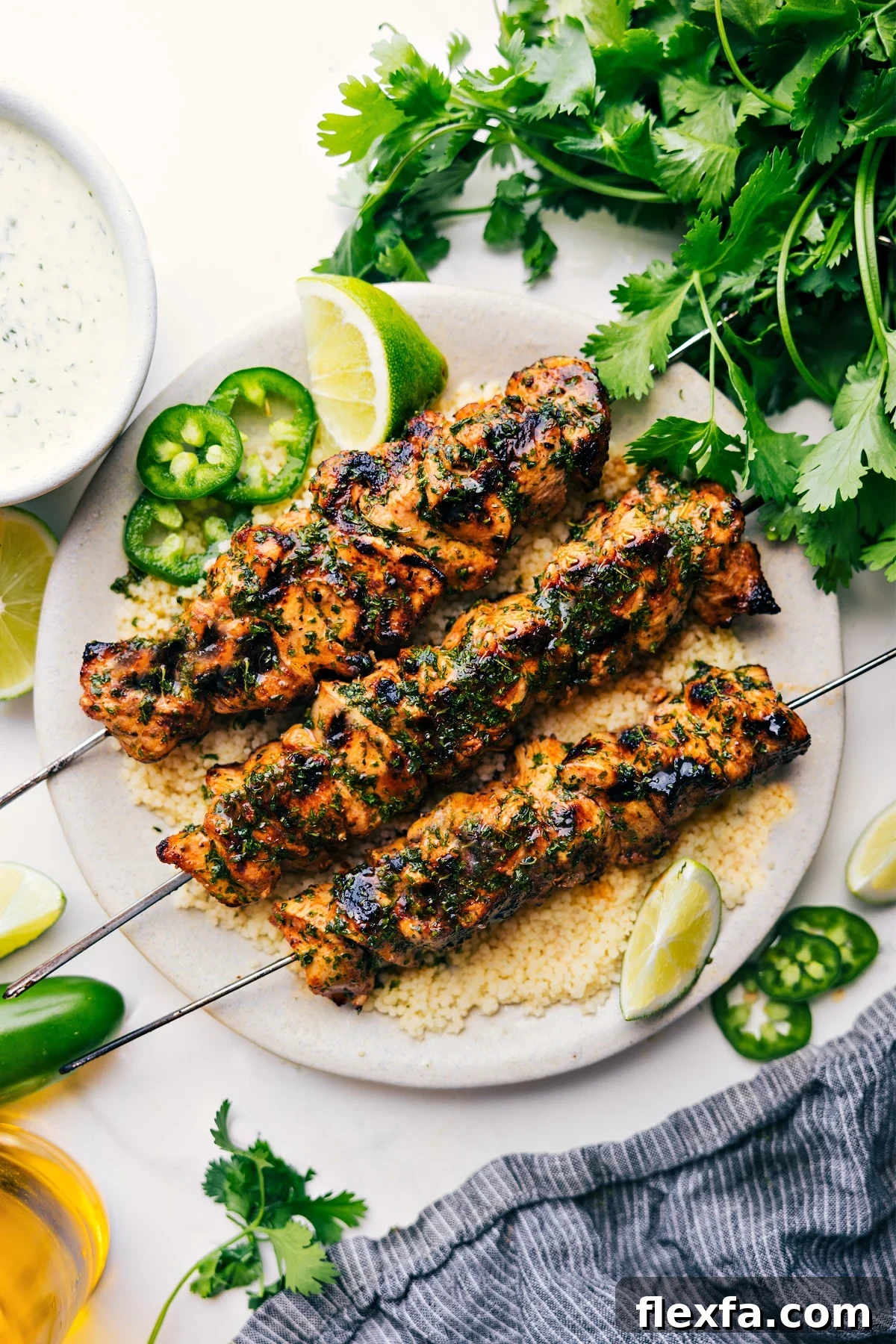 Chicken skewers served over fluffy couscous on a plate with lime wedges, sliced jalapeños, and creamy green sauce on the side.